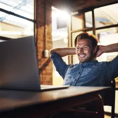 Uśmiechnięty mężczyzna w back office, z rękami za głową, patrzy na laptopa.
