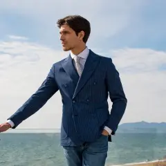 Uomo con giacca blu a doppio petto a righe, camicia bianca e cravatta, appoggiato a una ringhiera con vista mare.