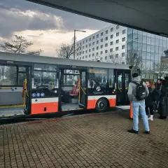 Ludzie czekają na autobus, który może być odpowiedzią na pytanie "jak dojechać do Pragi". W tle nowoczesny budynek i niebo.