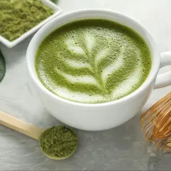 Zielona matcha w filiżance z pianką, obok miarka z proszkiem i bambusowa miotełka. Czy matcha ma kofeinę? Tak, zawiera ją!