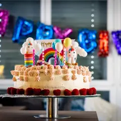 Kolorowy tort z jednorożcami i tęczą, idealny na zabawy na urodziny 7 latka. Balony i napis "Happy Birthday" w tle.
