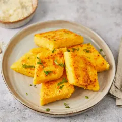 Gebratene Polenta-Ecken, italienisch garniert mit Petersilie, auf einem Teller. Daneben eine Reibe mit Parmesan.