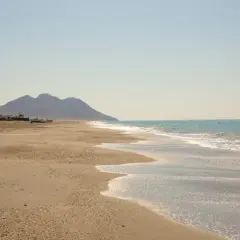 La playa de San Miguel, con sus aguas turquesas y arena dorada, se extiende hacia unas montañas. En la orilla, barcos y una torre antigua esperan la marea.