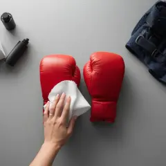 A hand wipes red boxing gloves with a white cloth. Beside them are cleaning supplies and a folded towel with a navy jacket. Don't put boxing gloves in the washing machine; clean them by hand.