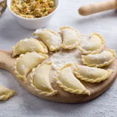 Gotowe do gotowania pierogi ruskie, ułożone na desce. Obok miska z farszem, mąka i wałek. Idealny przepis na pierogi ruskie.