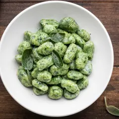 Gnocchi di spinaci freschi, conditi con foglie di salvia, in una ciotola bianca su un tavolo di legno scuro.