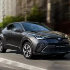 Ciemnoszary Toyota C-HR Hybrid w ruchu miejskim, z rozmytym tłem budynków i drzew.
