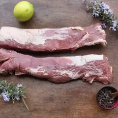 Deux pièces de filet mignon cru sur une planche en bois, accompagnées de romarin fleuri, d'une lime et de baies poivrées.