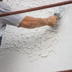 Un operaio applica intonaco rustico con una spatola, creando una texture grezza e irregolare su una parete esterna.