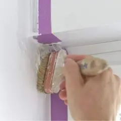 Hand streicht mit Pinsel und Klebeband eine weiße Wand.