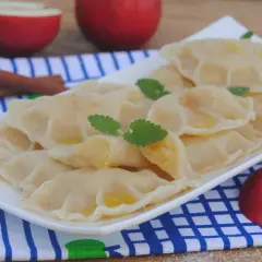 Pyszne pierogi z jabłkami, posypane cynamonem, z listkami mięty i kawałkami jabłek w tle.