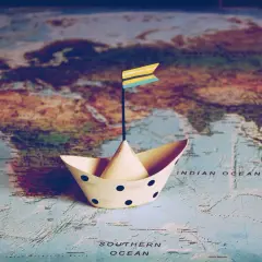 Papierowy stateczek z flagą gotowy do wyprawy dookoła świata. Mapa świata pod nim pokazuje kontynenty i oceany.