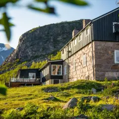 STF Kebnekaise fjällstation badar i solljus. En stenbyggnad med mörka trädetaljer reser sig mot en dramatisk bergsklippa.