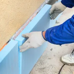 Pracownik w białych rękawiczkach układa niebieski styropian przy ocieplanej ścianie. To kluczowy etap w rankingu styropianu dla izolacji.