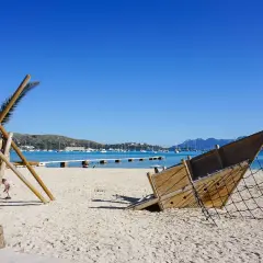 Spielplatz am Strand im mallorcas norden. Ein hölzernes Schiffswrack und eine Kletterstruktur stehen im Sand, Boote liegen im türkisfarbenen Wasser.