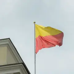 Flaga żółto czerwona powiewa na maszcie obok zabytkowego budynku.