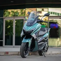 Scooter onex motos color verde menta, con parabrisas alto, estacionada frente a un edificio moderno con puertas de cristal.