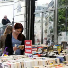 Une jeune femme choisit un livre dans une librairie en plein air, devant une vitrine reflétant des bâtiments parisiens. Une sélection de livres, dont des romans policiers et de suspense, est exposée, rappelant une maison d'édition jeunesse.