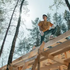 Młody mężczyzna w białych rękawiczkach stoi na konstrukcji domku drewnianego, otoczony lasem sosen.