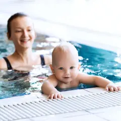 Uśmiechnięty niemowlak w basenie, trzymający się brzegu. Mama w tle obserwuje go z radością.