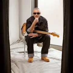 Alex Britti, con occhiali da sole e chitarra, sembra pensieroso, quasi gelido, mentre riflette sui suoi testi.