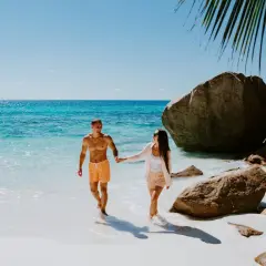 Para idealny czas na podróż na Seszele, para spaceruje po rajskiej plaży z turkusową wodą i skałami.
