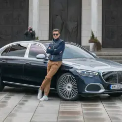 Elegancki mężczyzna opiera się o lśniącego Maybacha S580 przed monumentalnym budynkiem.