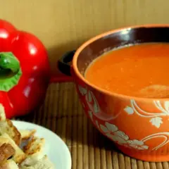 Aksamitny zupa krem z papryki, podana z chrupiącymi grzankami. Obok leży świeża, czerwona papryka.