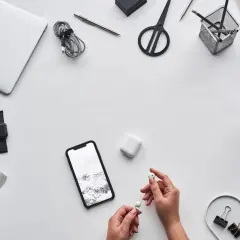 Ręce trzymające słuchawki AirPods obok telefonu z Androidem, laptopa i kawy. Czy AirPods działają z Androidem? Tak!