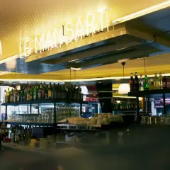 Bar lumineux avec enseigne néon "LE MANSART PARIS". Bouteilles d'alcool et verres alignés sur les étagères.