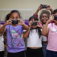Five girls hold up old cameras, learning what to do with them by taking pictures.