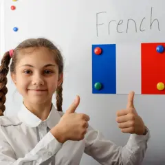 Uśmiechnięta dziewczynka z warkoczami pokazuje kciuki w górę. Na tablicy napis "French" i flaga Francji. Edukacja we Francji ciekawostki.