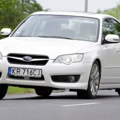 Białe Subaru Legacy z polskimi tablicami KR 716CJ jedzie po drodze wśród zieleni.