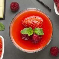 Un bicchiere di coulis di fragole con fragola, lamponi e menta. Accanto, fragole fresche, cioccolato bianco, latte e un baccello di vaniglia.