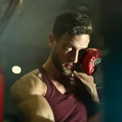 A boxer with a beard and intense blue eyes, wearing a red tank top and a red boxing glove, poses in a gym. He's ready to jump into action.