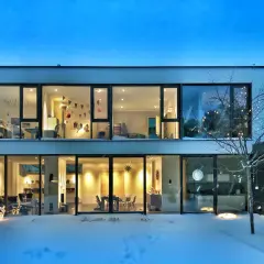 Casa moderna con grandes ventanales iluminados, mostrando interiores acogedores. La nieve cubre el exterior, y luces navideñas decoran un árbol. ¡2500 lumens cuanto ilumina!