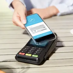 Mit handy bezahlen: Eine Hand hält ein Smartphone über ein Kassenterminal, um kontaktlos zu bezahlen.