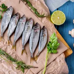 Seis jureles pescado frescos, listos para cocinar, acompañados de perejil, romero, limón y champiñones sobre papel de estraza y tabla de madera.