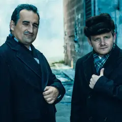 Obsada filmu "Gierek": dwaj mężczyźni w płaszczach i szalikach, jeden w futrzanej czapce, stoją obok czarnego samochodu.