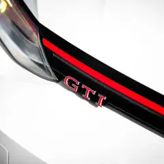Detalle de un coche blanco con las siglas GTI en rojo y cromo, sobre una franja negra y roja.