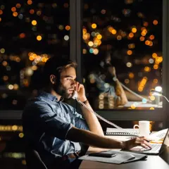 Un homme travaille tard, au téléphone pour une réclamation AXA, devant un paysage urbain nocturne illuminé.