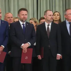 Mężczyźni w garniturach, być może posłowie, stoją w rzędzie, trzymając czerwone dokumenty.