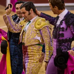 Tres toreros con trajes de luces, uno con coleta de torero, son felicitados en la arena.