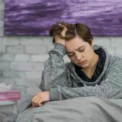 Müde Person im Bett, die sich an den Kopf fasst. Mögliche Anzeichen von Blutkrebs wie Müdigkeit und Kopfschmerzen sollten ernst genommen werden.