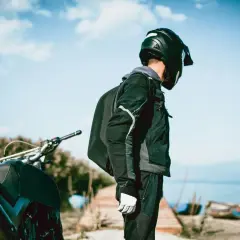 Motociclista con casco negro, chaqueta y guantes blancos, junto a su moto. El tejido de poliéster 600d vs Cordura es ideal para su aventura.