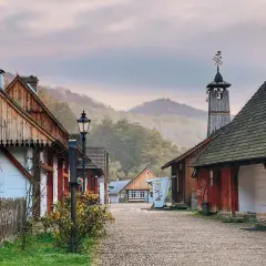 Sanok skansen: urokliwa, brukowana uliczka z drewnianymi domami i wieżą z wiatrowskazem, otoczona mglistymi wzgórzami.