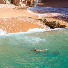 Eine Frau schwimmt im türkisfarbenen Wasser vor einem goldenen costa brava strand, umgeben von Felsen.