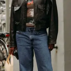 Personne portant une veste en cuir noire, un haut à paillettes et un jean bleu, tenant un sac beige.