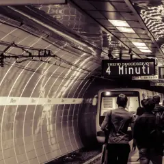 Persone attendono il treno sulla metro linea b. Un cartello indica "Treno successivo: 4 minuti".