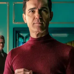 Berlin z "La Casa de Papel" w bordowym golfie, zamyślony. W tle inny mężczyzna.